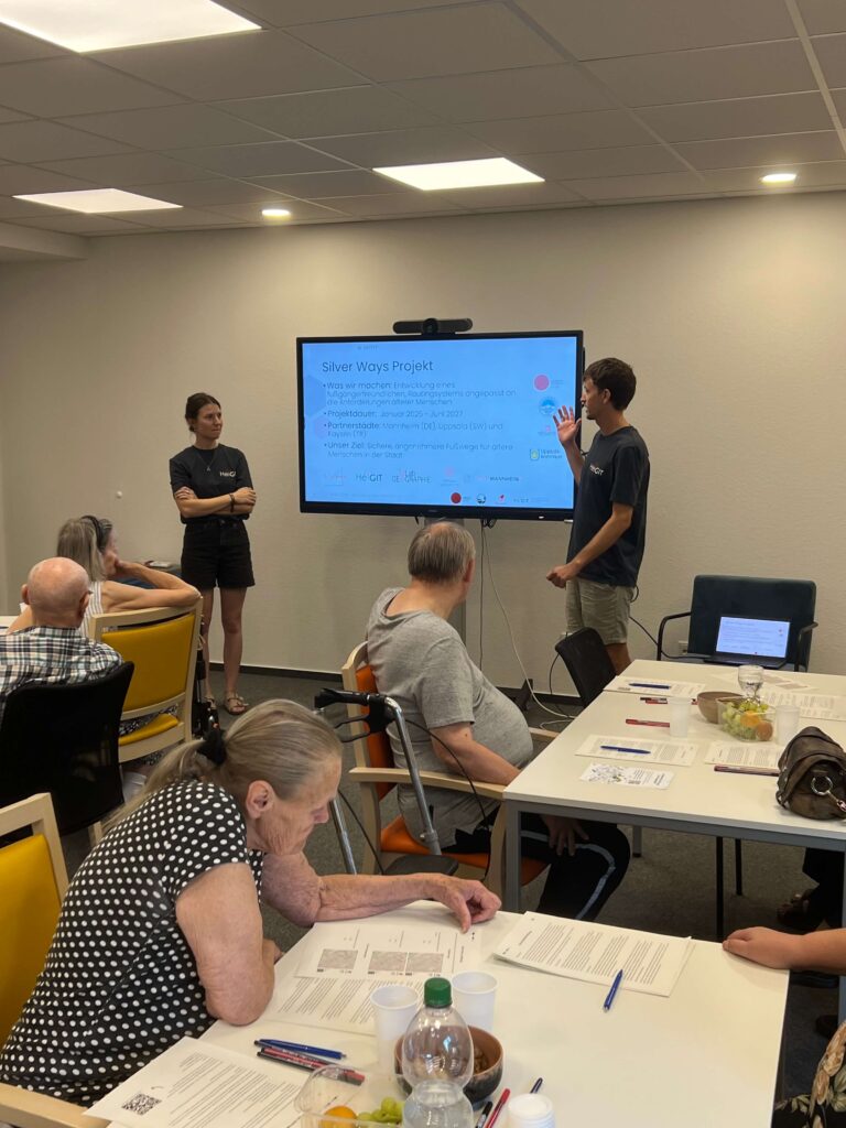 Person standing and pointing at a screen displaying a presentation titled 'Silver Ways Projekt' with bullet points, another person standing with arms crossed, and seated audience members at tables with papers and bottles